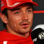 Charles Leclerc wearing a red Ferrari cap and team hoodie, speaking into an FIA Formula 1 microphone during Thursday's press conference at the 2026 Mexico City Grand Prix.