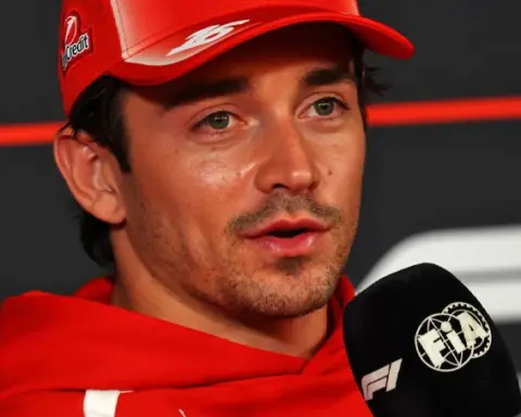 Charles Leclerc wearing a red Ferrari cap and team hoodie, speaking into an FIA Formula 1 microphone during Thursday's press conference at the 2026 Mexico City Grand Prix.