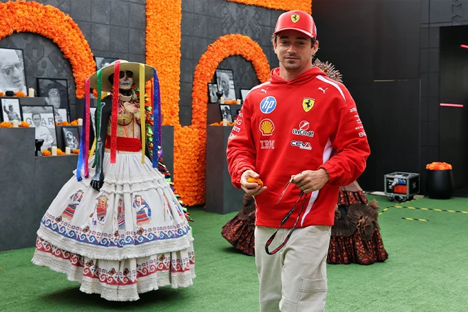 Charles Leclerc wearing Ferrari team clothing and a red cap beside a traditional Día de los Muertos display featuring a decorated skeleton and orange marigolds in Mexico City.